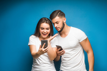 Naklejka premium Portrait of curious young couple man and woman in basic t-shirts smiling while peeking at each other cellphones isolated over background.