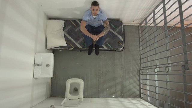 Male Prisoner in Prison Cell. The inmate is sat on his bed. Aerial view from above