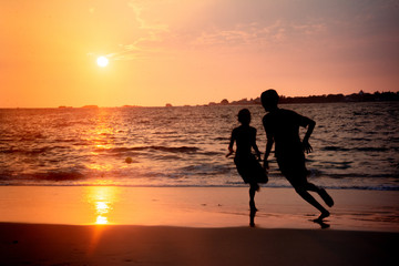 Las siluetas de dos niós corren por la orilla de una playa al atardecer, con el sol de fondo