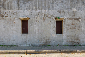 Front side sunny view of concrete sidewalk and old rough scratch concrete wall with small wooden two windows in old town of Bangkok, Thailand.