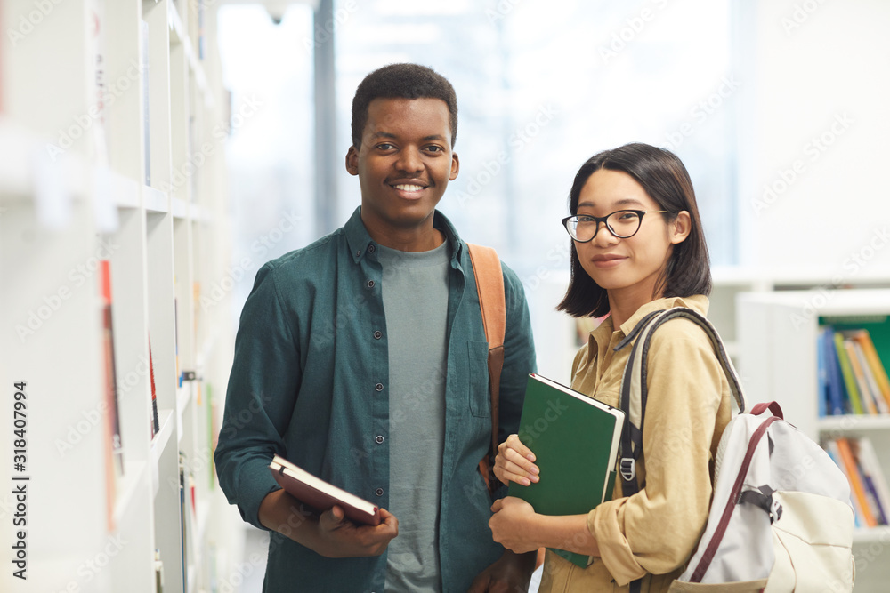 Canvas Prints waist up portrait of two international students looking at camera and smiling while posing by shelve