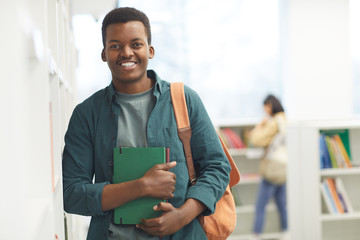 Waist up portrait of African-American student looking at camera and smiling while posing by shelves in college library, copy space