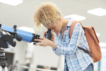 Side view portrait of funky female student looking into telescope in college library, copy space © Seventyfour