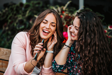 Two cheerful women listening music with cellphone and earphones and singing while sitting outdoor