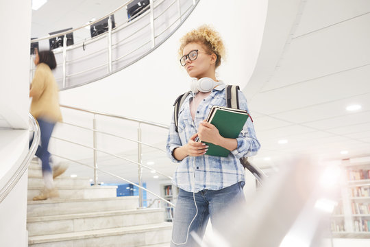 Waist up portrait of female student holding books while going down spiral stairs in modern college interior, copy space
