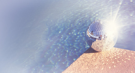 Party pool summer concept image with disco ball beside water edge and rays of sun glowing on globe.  Copy space and banner horizontal sizing.