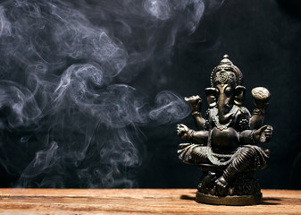 Hindu god Ganesha on black background. Statue on wooden table with a smoke of incense. Copy space.