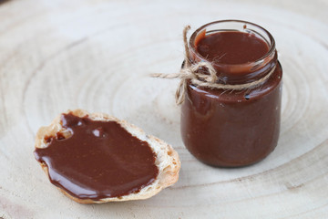 Open glass jar with some chocolate paste, chocolate spread on bread on light wooden background. Side view, copy space