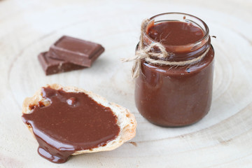 Open glass jar with some chocolate paste, chocolate spread on bread, two pieces of dark chocolate on wooden background.