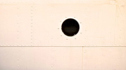 Riveted steel plates of a white topside of a classic ship with single round portlight window.