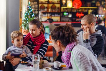 Fototapeta na wymiar Family at cafe or restaurant sitting by the table small boy child holding a smart phone while the aunt woman is talking to him in mothers lap sitting caucasian