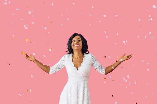 Portrait of a happy young woman in white dress and black hair smiling while celebrating with arms out with confetti falling