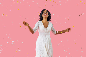 Obraz na płótnie Canvas Portrait of a happy young woman in white dress and black hair smiling while dancing with arms out and confetti falling