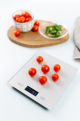 Red cherry tomatoes on gray digital kitchen scales. Bowl with tomatoes and plate with fresh green basil on wooden board on background. Selective focus. Kitchen equipment concept. Healthy eating.
