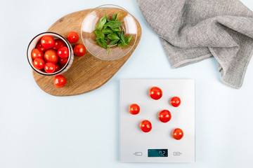 Red cherry tomatoes on gray digital kitchen scales. Near bowl with tomatoes, plate with fresh green basil on wooden board and towel. Close up, top view. Kitchen equipment concept. Weighing products.