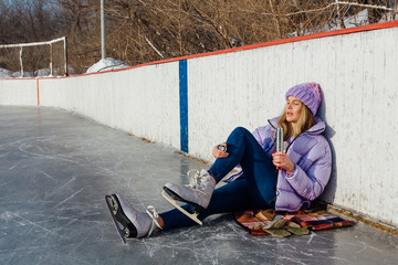 Lovely young woman relaxing after riding ice skates and drinking hot drink from termo pot on the ice rink.