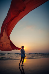 Young woman walks along sandy beach of the sea, holding red long cloth in the wind against the background of setting sun. Freedom and vacation concept. Photo from the back, no face.