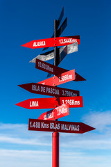 Red crossroad signpost showing the distance of several places on earth with a partially cloudy blue sky in the background. Alaska, Falklands, Lima, Sao Paulo.