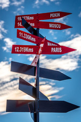 Red crossroad signpost showing the distance of several places on earth with a partially cloudy blue sky in the background. Pekin, Moscow, Paris, Madrid.