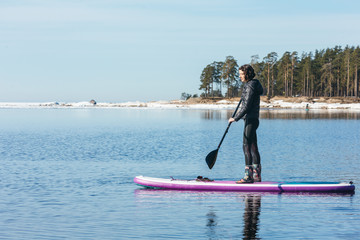 Fototapeta na wymiar Attractive caucasian woman in black wetsuit paddle on SUP board with an oar. Female floating on stand up paddle board in the sea. In the background trees. Winter season and active leisure concept.