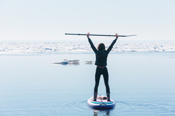 Slim woman in wetsuit paddle on SUP board and holds oar over her head. Female floating on stand up paddle board in the sea. Active leisure concept. An attack of happiness and euphoria.