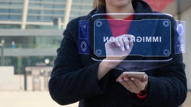 Unrecognizable woman interacts HUD hologram with text Immigration. Girl in the coat uses the technology of the future mobile screen on the background of the city