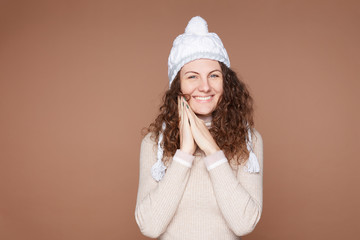 Fototapeta premium Pretty happy European woman dressed in cozy outfit, keeps hands on cheeks, wears white knitted hat, cashmere sweater, poses on beige studio wall. Cheerful lovely feminine lady feels amused, isolated.