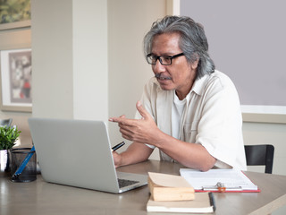 Asian Senior man wear eyeglasses is using a laptop and thinking while sitting on chair in room at home. Old elderly male Technology communication. Business after retirement concept.