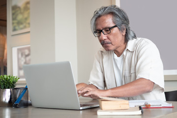 Fototapeta premium Asian Senior man wear eyeglasses is using a laptop while sitting on chair in room at home. Old elderly male Technology communication. Business after retirement concept.
