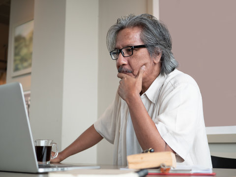 Asian Senior Man Wear Eyeglasses Is Using A Laptop And Thinking While Sitting On Chair In Room At Home. Old Elderly Male Technology Communication. Business After Retirement Concept.