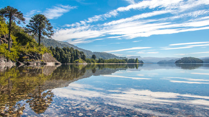 hermoso paisaje de la patagonia, un lago bordeada por un cerro y bosque de araucarias con cielo azul y nubes reflej&aacute;ndose en el agua