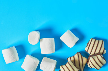 Scattered striped white and dark chocolate candies in shape of star and marshmallow in form of cylinder lies on blue countertop. Space for text. Top view
