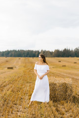 Fototapeta na wymiar A fair haired young woman farmer in a white dress in a field with stacks of straw enjoys a summer day