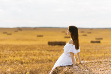 A fair haired young woman farmer in a white dress in a field with stacks of straw enjoys a summer day