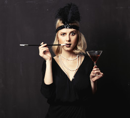 Beautiful blonde woman in twenties years clothes with smoking pipe and cocktail on dark background