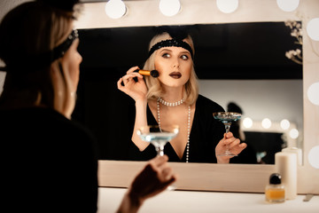 Beautiful blonde woman in twenties years clothes applying makeup near a mirror with bulbs