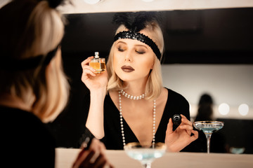 Beautiful blonde woman in twenties years clothes applying makeup near a mirror with bulbs