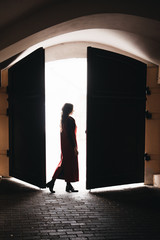 Fototapeta premium Outdoor portrait of a stylish young girl on the street of an old city in black clothes and a long red vest. Silhouette photo. A model poses on the street of a European city