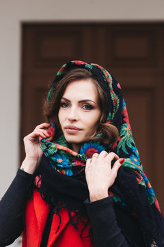 Outdoor portrait of a stylish young girl on the street of an old city in black clothes and a long red vest and a Russian scarf on her head. A model poses on the street of a European city