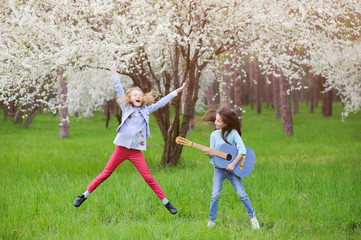 ecstatic live outdoor rock music concert by two little young girls jumping singing and playing guitar in spring blossom green park with white flowering tree