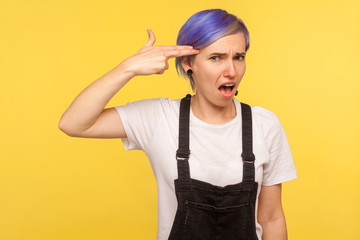 Portrait of depressed frustrated hipster woman with violet short hair in denim overalls pointing finger gun to head, looking bored tired of life, pissed off. isolated on yellow background, studio shot