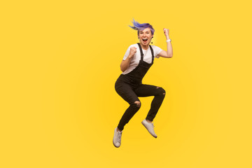 Fototapeta na wymiar Yes i did it! Portrait of enthusiastic lively hipster girl with violet hair in overalls jumping in air with raised fists, showing yes i did it gesture, life energy. yellow background, studio shot
