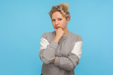 Smart and thoughtful girl with short curly hair in sweatshirt holding her chin and pondering idea, making difficult decision, looking uncertain doubtful. indoor studio shot isolated on blue background