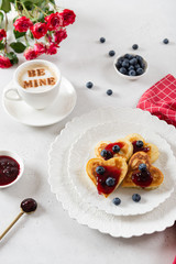 Homemade heart shaped pancakes with cherry jam and blueberries on white porcelain plate and a cup of cappuccino with message be mine.