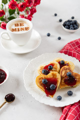 Valentine pancakes in heart shape with jam and berries on white plate with a cup of coffee and roses bouquet. St. Valentines Day surprise, breakfast concept.
