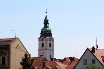 Church bell tower with analogue clocks on all sides and large metal cross on top covered with green metal tiles rising high above city family houses rooftops on clear blue sky background