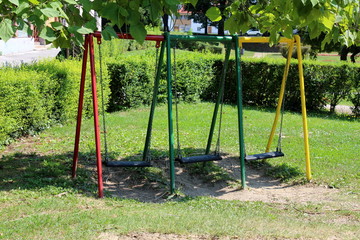 Fototapeta premium Colorful outdoor public playground equipment metal swing with three plastic seats in shade of tall old tree surrounded with grass and hedge in local public park