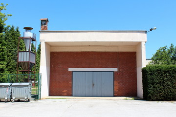 Large red bricks garage with elongated garage doors made from narrow wooden boards with faded color next to small storage silo surrounded with dense hedge and trees on clear blue sky background