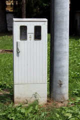 Narrow small locked grey plastic electrical meter and fuse box next to tall concrete utility pole in suburban family house backyard surrounded with uncut grass