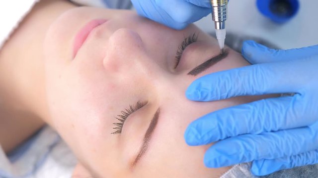 Cosmetologist making eyebrows microblading procedure in beauty salon for girl using tattoo apparatus, face closeup. Beautician in gloves is doing permanent makeup. Beauty industry concept.
